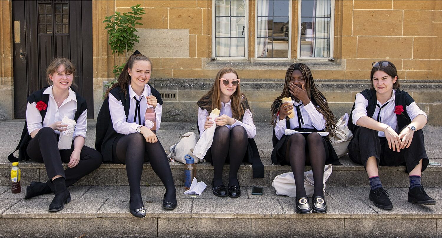 5 female students in their academic gowns sat on steps outside with each one holding either a drink or a sandwich in hand