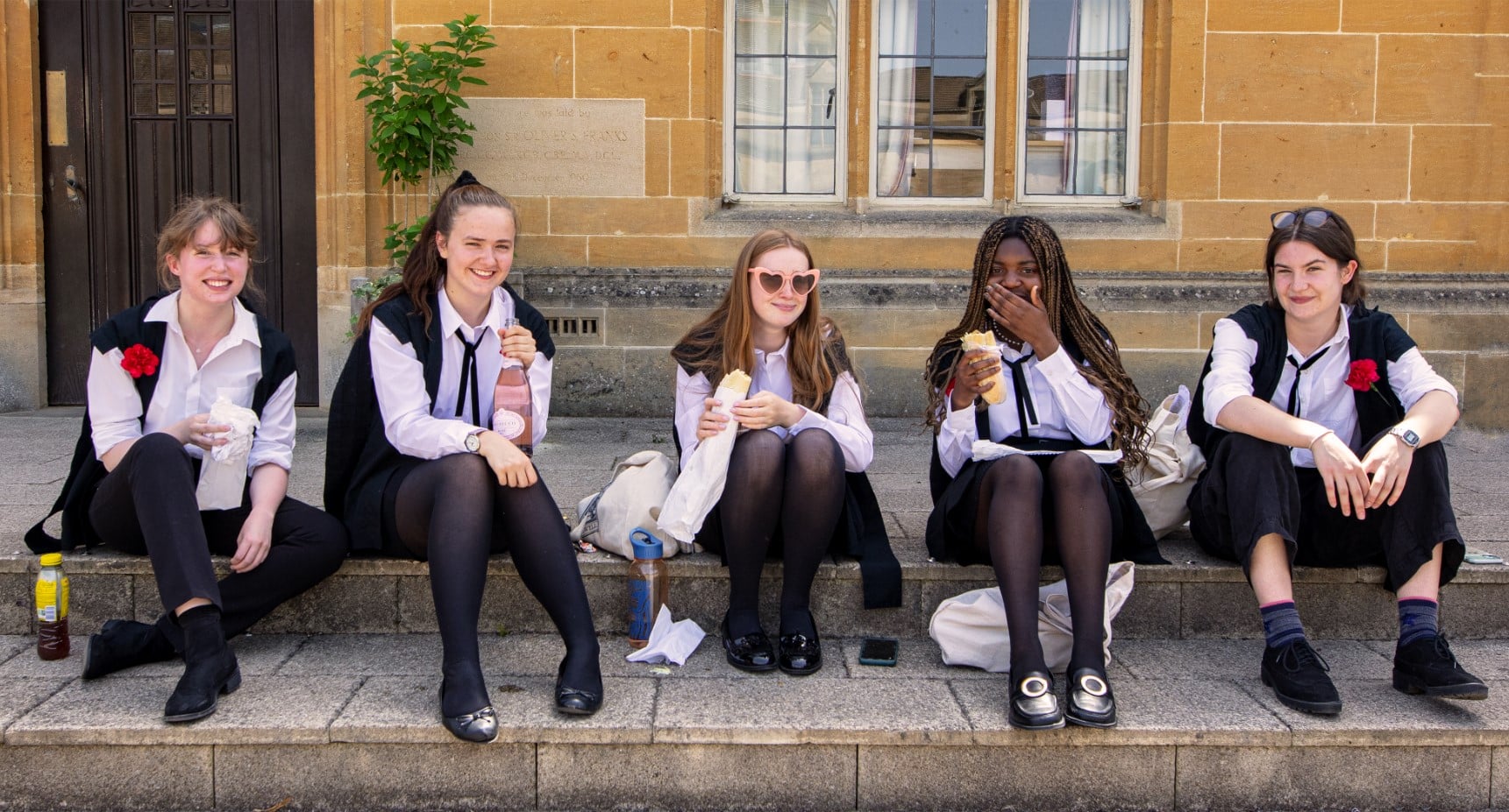 5 female students in their academic gowns sat on steps outside with each having a drink or sandwich in hand