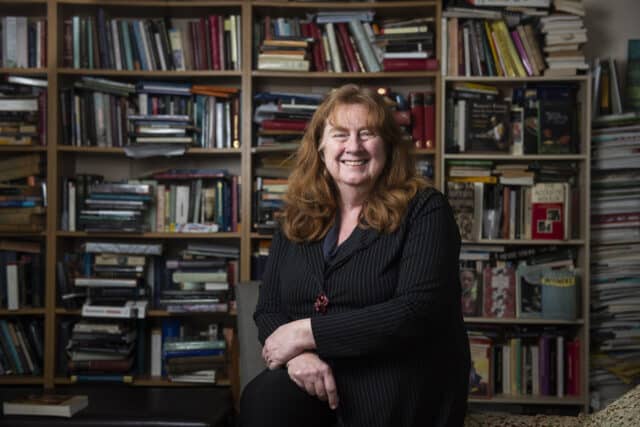 Photo of Ros with her hands crossed and rested on her lap and bookshelves in the background