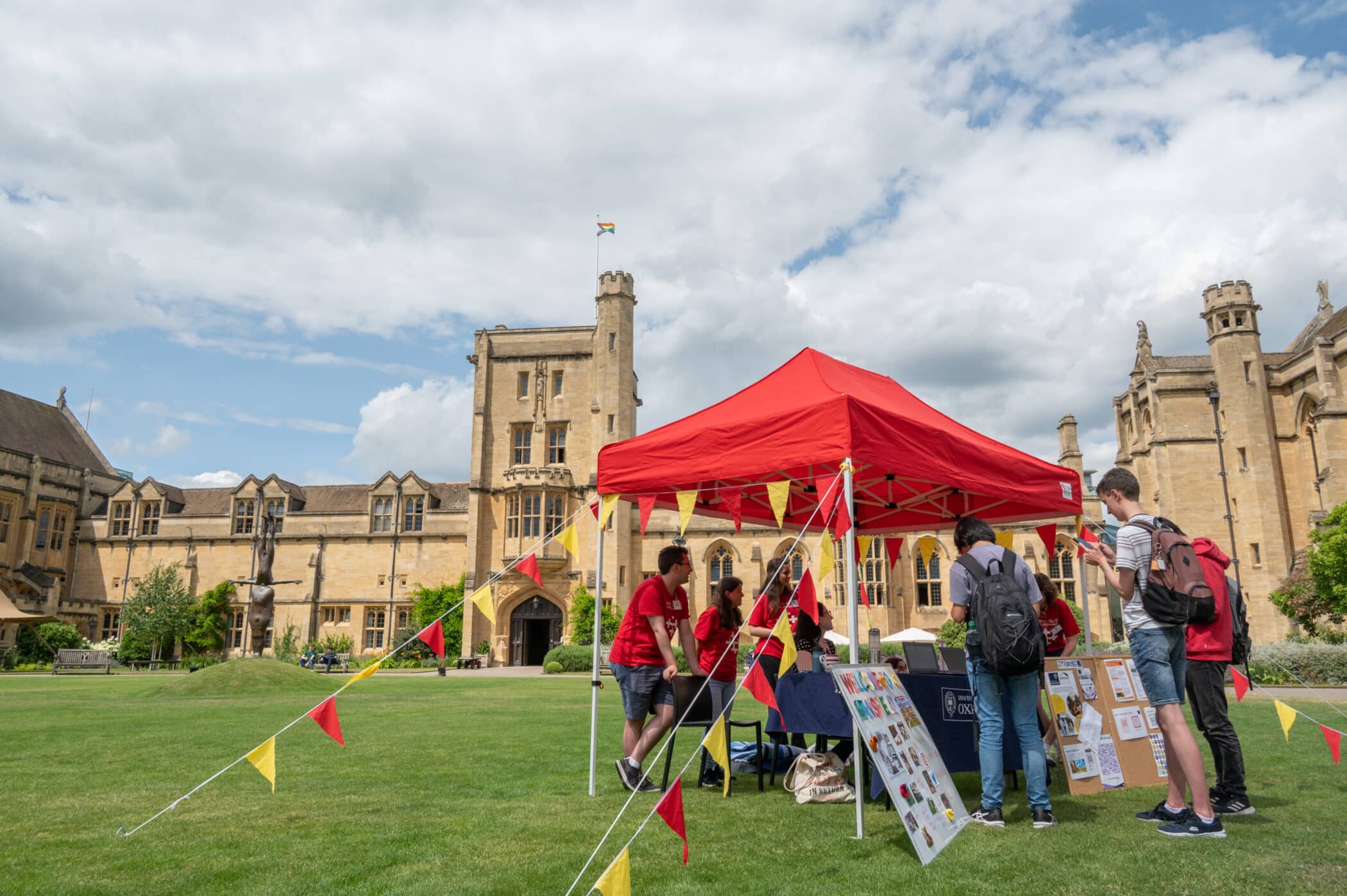 Photograph of the June 2023 Open Day. A red gazebo and yellow and red bunting surrounded by people. Behind is Mansfield's main building.